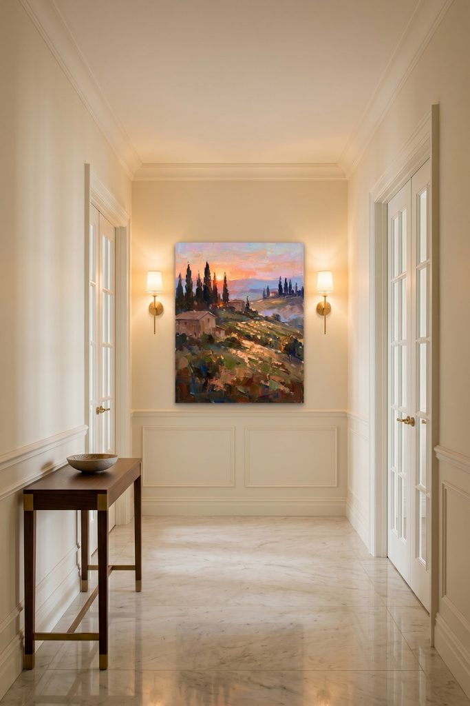 A warm Tuscan sunset landscape painting displayed in an elegant bright hallway with classic white wainscoting and warm wall sconces.